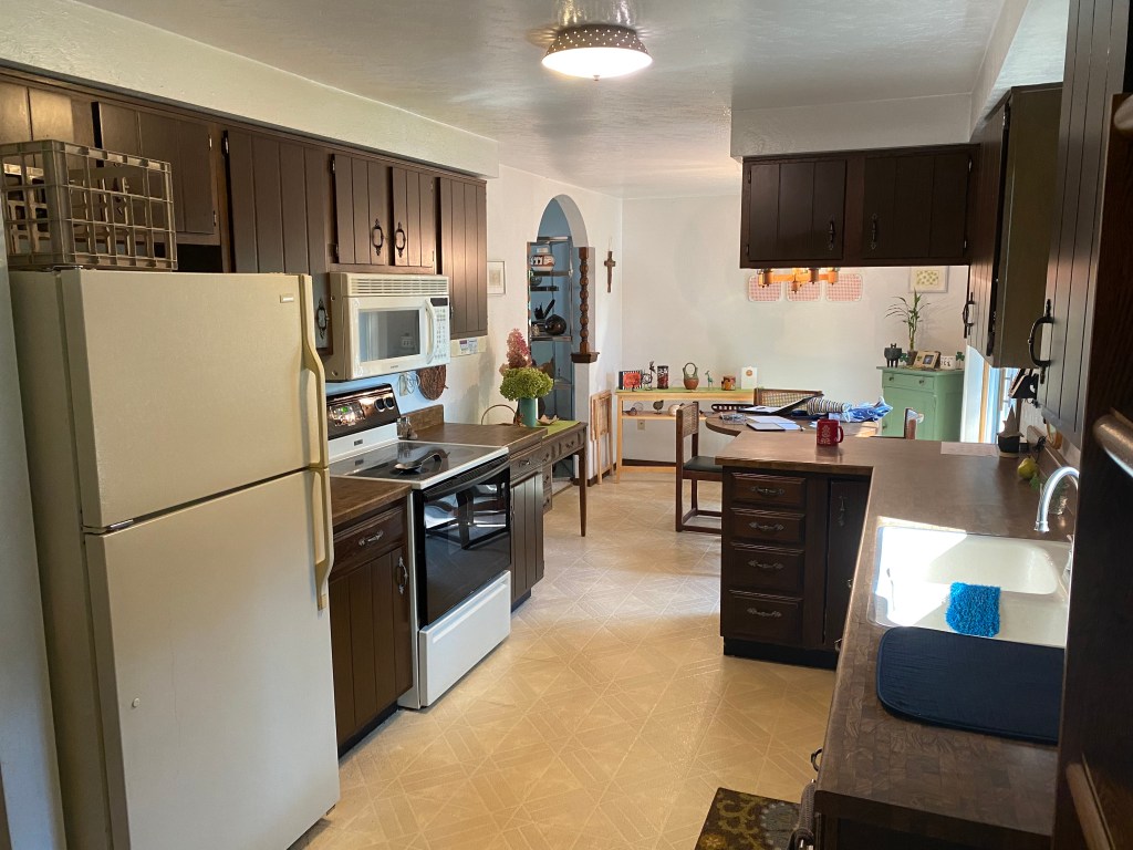 Before image of kitchen standing in the corner looking across the room at the fridge, stove, and outdated cabinets